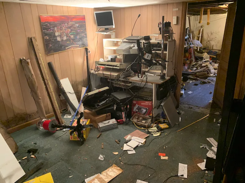 A cluttered, e-waste-filled basement office in North NJ, featuring old computers, monitors, a stereo receiver, and mountains of tangled cables before professional e-waste removal by Junk Doctors NJ.
